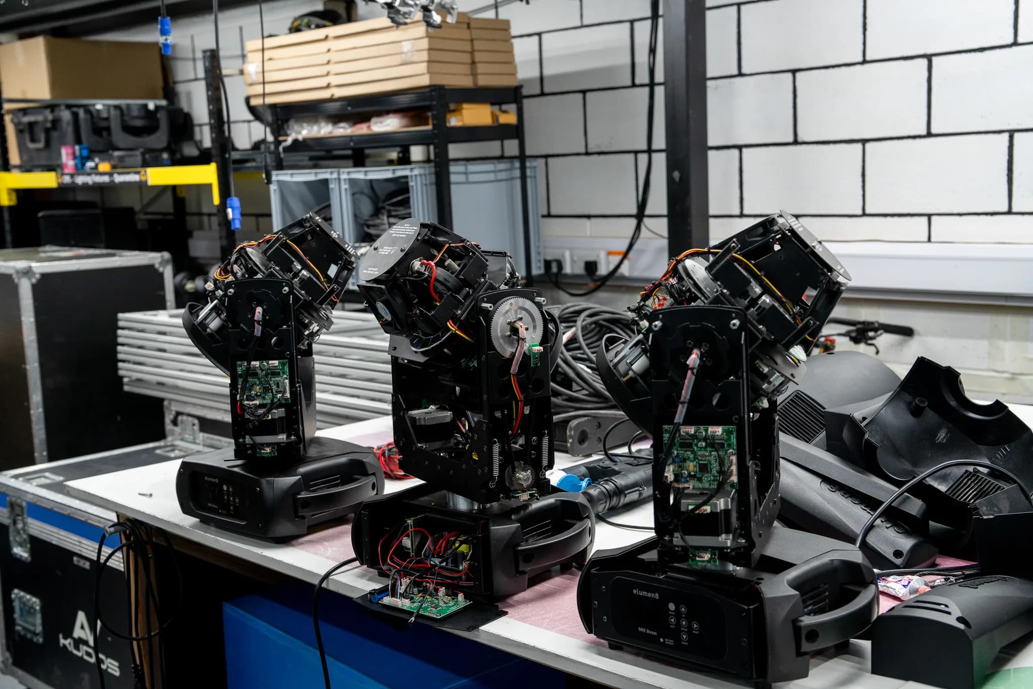 Three disassembled moving head lights open for repair on a workbench, exposing internal circuit boards and wiring.