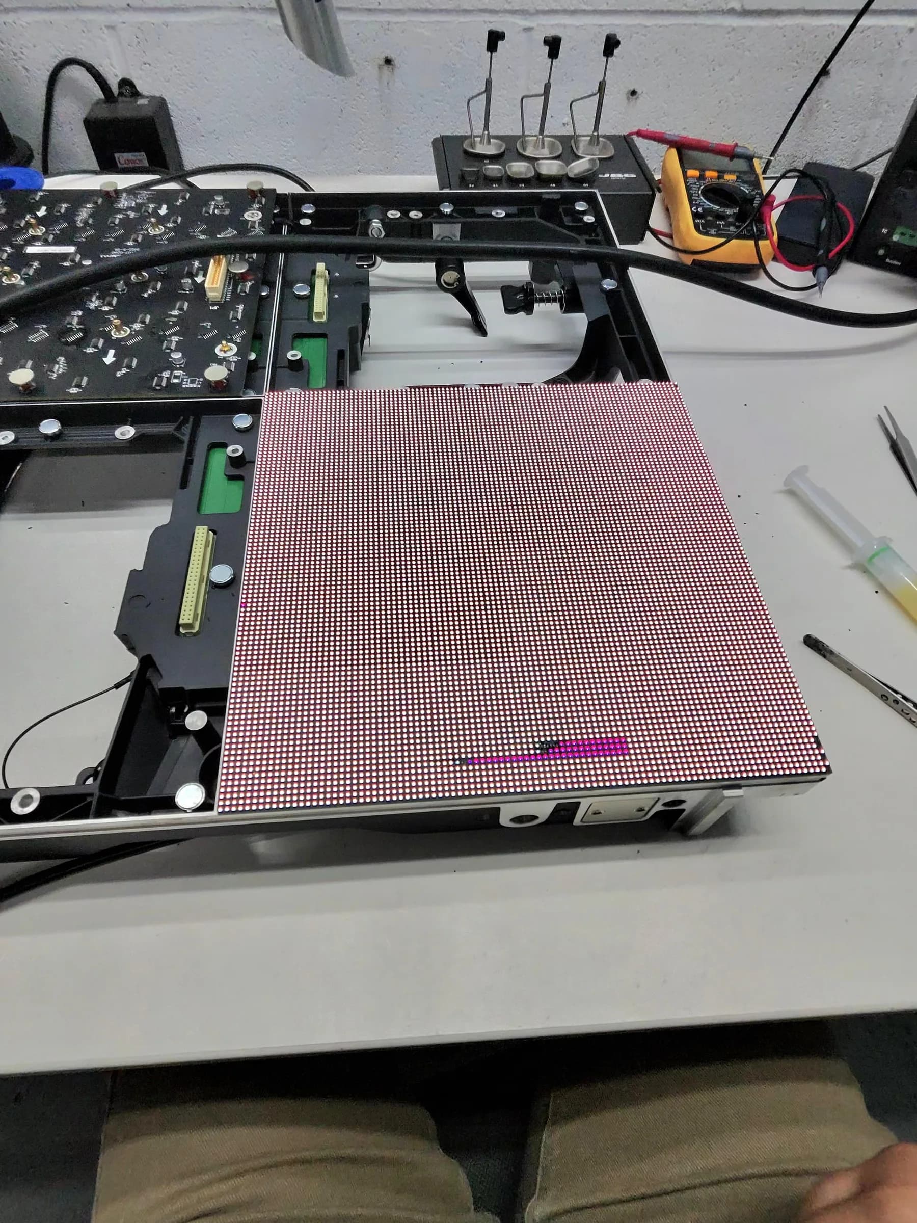 LED display module on a repair bench showing a partially lit pixel test pattern near a multimeter and soldering station.