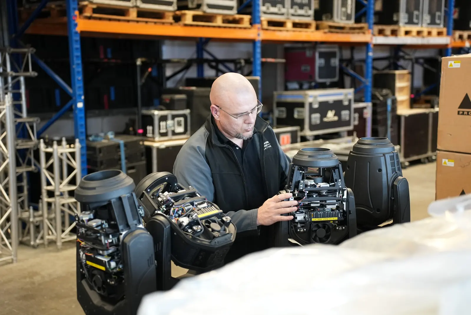 Bald technician in glasses inspects the open internal components of moving head lighting fixtures in a warehouse