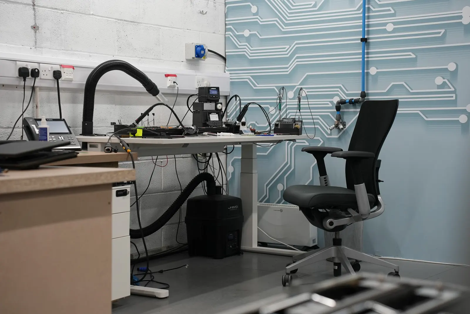 Electronics repair workbench with soldering station, fume extractor, and microscope in front of a circuit-patterned wall